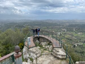 Hufeisenförmige Aussichtsplattform auf dem Kaiser's Throne, drei Menschen stehen darauf, eine Frau zeigt nach Westen. Im Hintergrund die Landschaft Korfus, der Himmel ist dunkelblau dicht bewölkt
