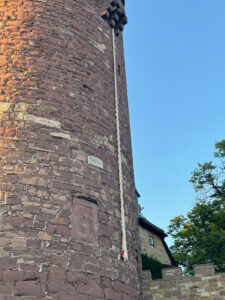 Wand eines runden Turms, aus einem Fenster oben hängt ein dickes, helles Tau als 'Rapunzelzopf' weit herunter