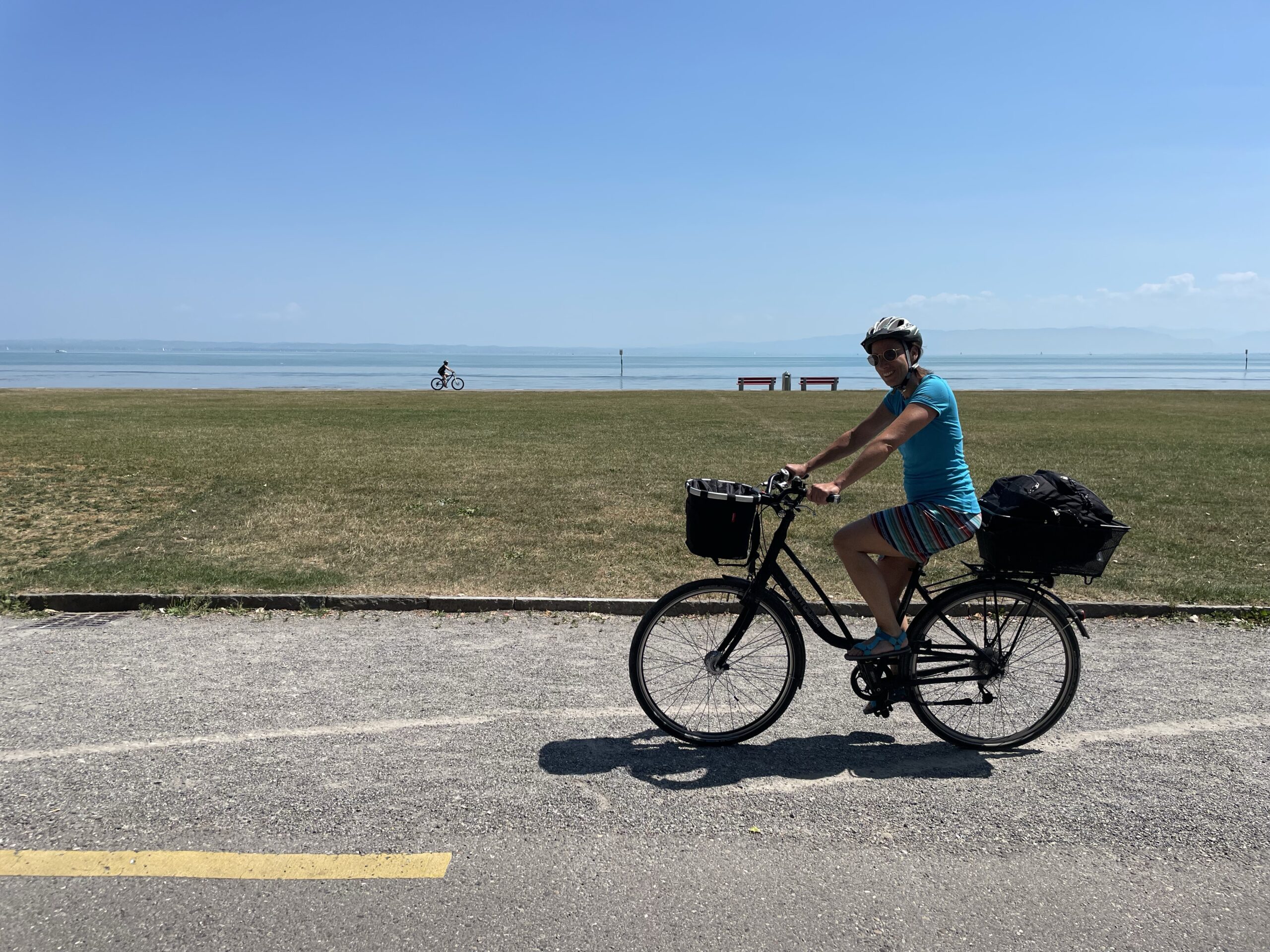 im Vordergrund eine Radlerin, die von rechts nach links fährt, dahinter ein großer, leerer Rasen. hinter dem Rasen eine Person auf einem Rad, die von links nach rechts fährt, dahinter der See und am Horizont ist die Sillouhette von Bergen gerade so noch zu erkennen