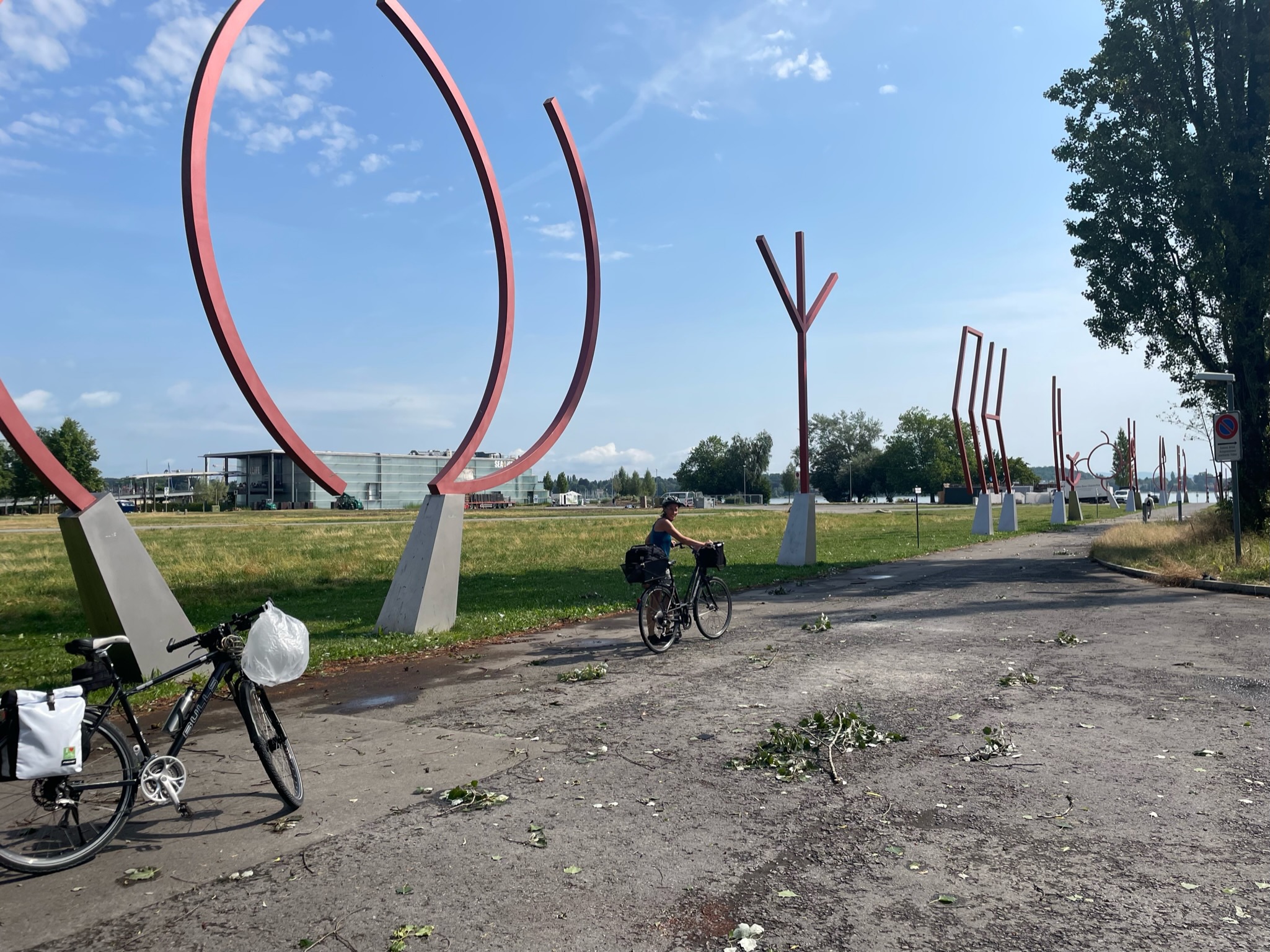 eine Reihe runenähnliche, etwa vier Meter hohe Skulpturen entlang eines geraden Weges, der auf den See zuführt. im Vordergrund ein Fahrrad und eine Radlerin, die ihr Fahrrad hält und zur Kamera zurückblickt