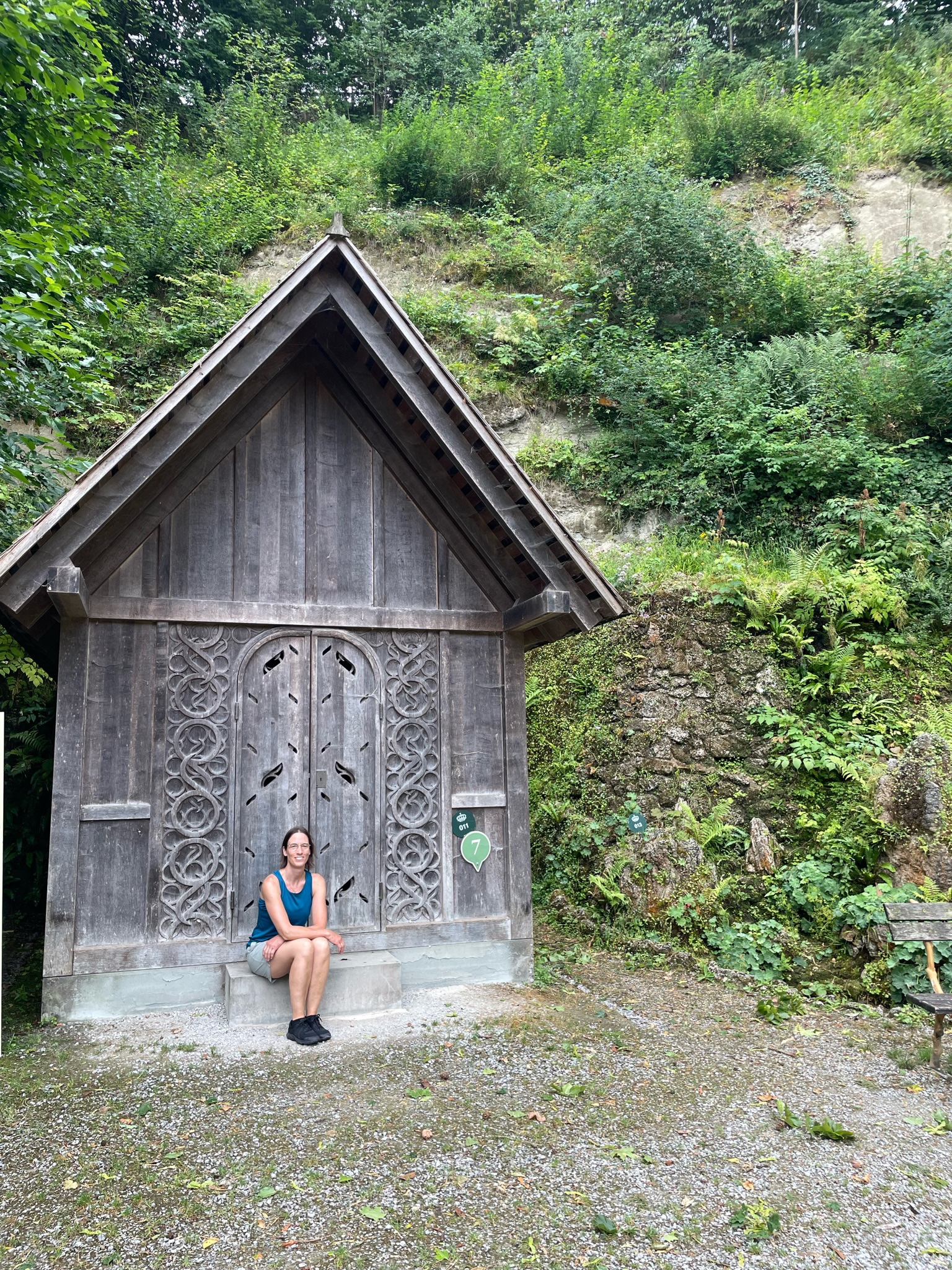 eine Frau sitzt auf der Eingangsstufe einer sehr schmalen, relativ hohen Hütte mit spitzem Dach. Die Hütte ist aus vergrautem, unbehandelten Holz gebaut, mittig ist eine kleine Rundbogentür (geschlossen) rechts und links davon sind ornamentale Schnitzereien angebracht. Direkt hinter der Hütte beginnt eine Felswand aus der Farn und einige Büsche wachsen