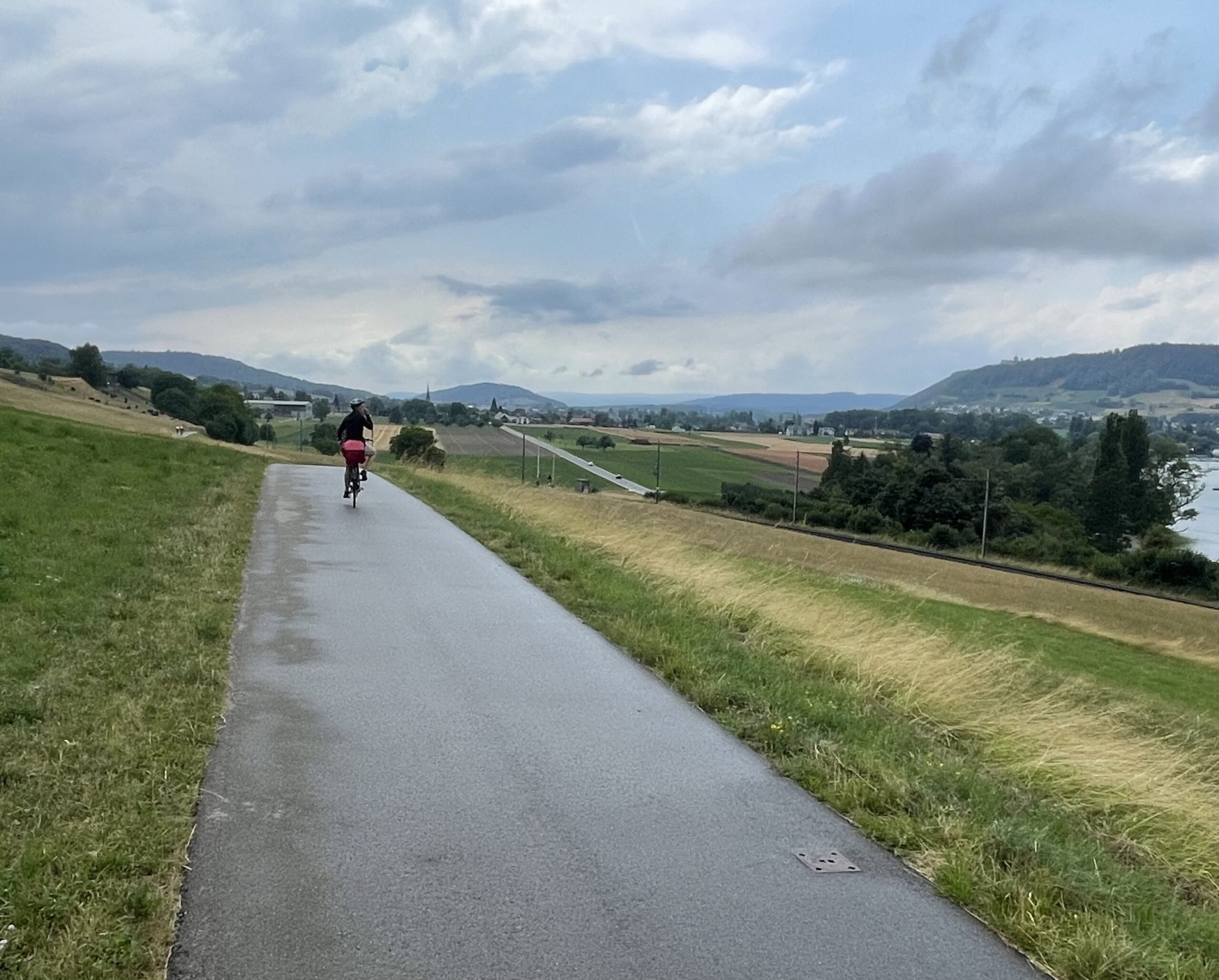 eine Radlerin, schon ein Stück entfernt auf einem glatten, asphaltierten Radweg entlang eines Hügels, rechts ist etwas tiefer der See hinter Bäumen zu sehen, Hügel mit Feldern, Wäldern und Hecken bestimmen eine vielgestaltige Landschaft