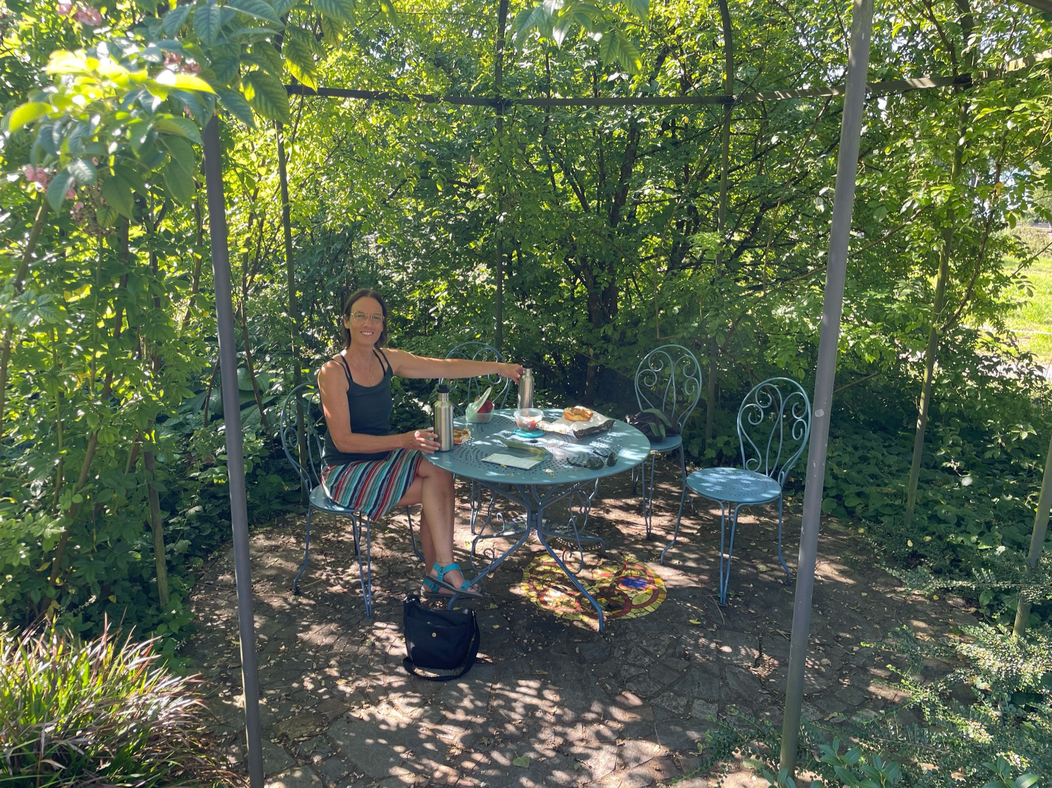 eine Frau in hellblau-rot-gestreiftem Rock, hellblauen Sandalen und dunkelblauem Top sitzt auf einem Stuhl einer hellblauen Metall-Sitzgruppe. Auf dem Tisch ist ein kleines Picknick aufgetischt, die Sitzgruppe steht im Grünen, unter einem Pavillongerüst, mit Büschen und Blumen im Hintergrund